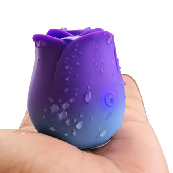 Close-up of a small gradient purple-to-blue silicone rose-shaped toy with water droplets, perched on a fingertip.